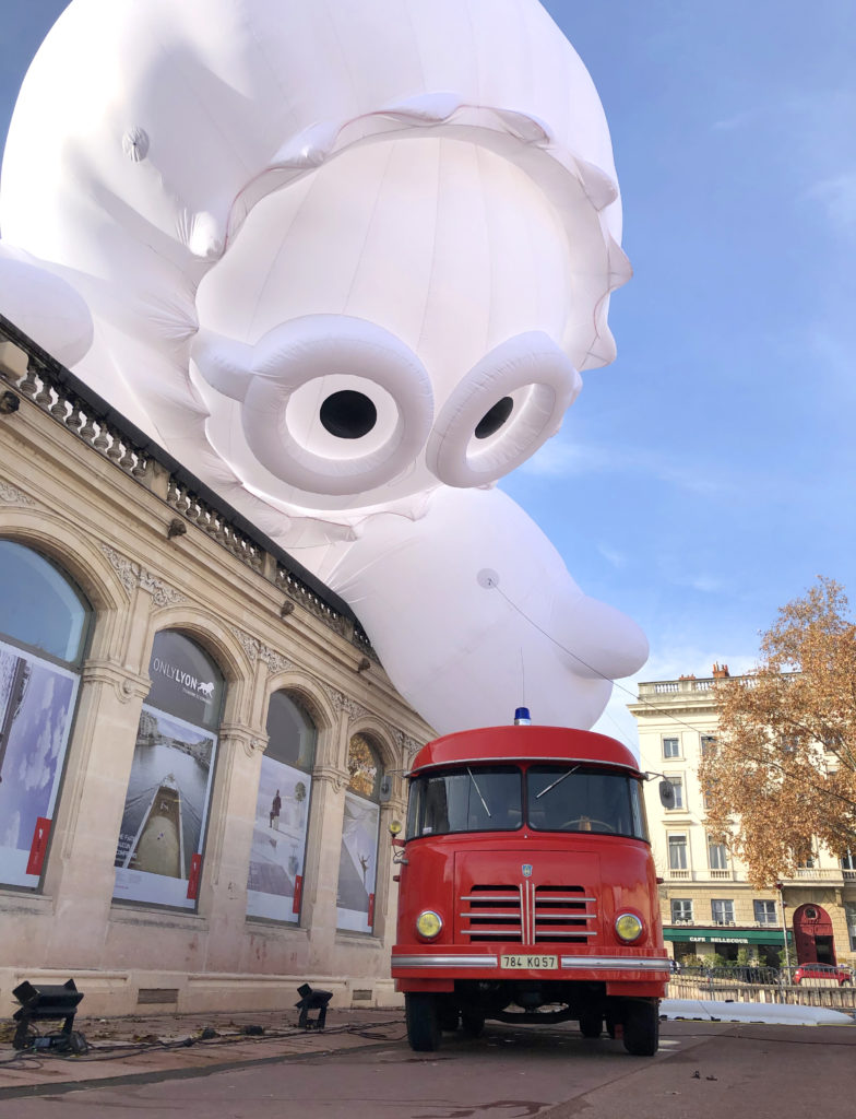 En pleine journée, un personnage gonflable joue avec un camion de pompier, place Bellecour à Lyon